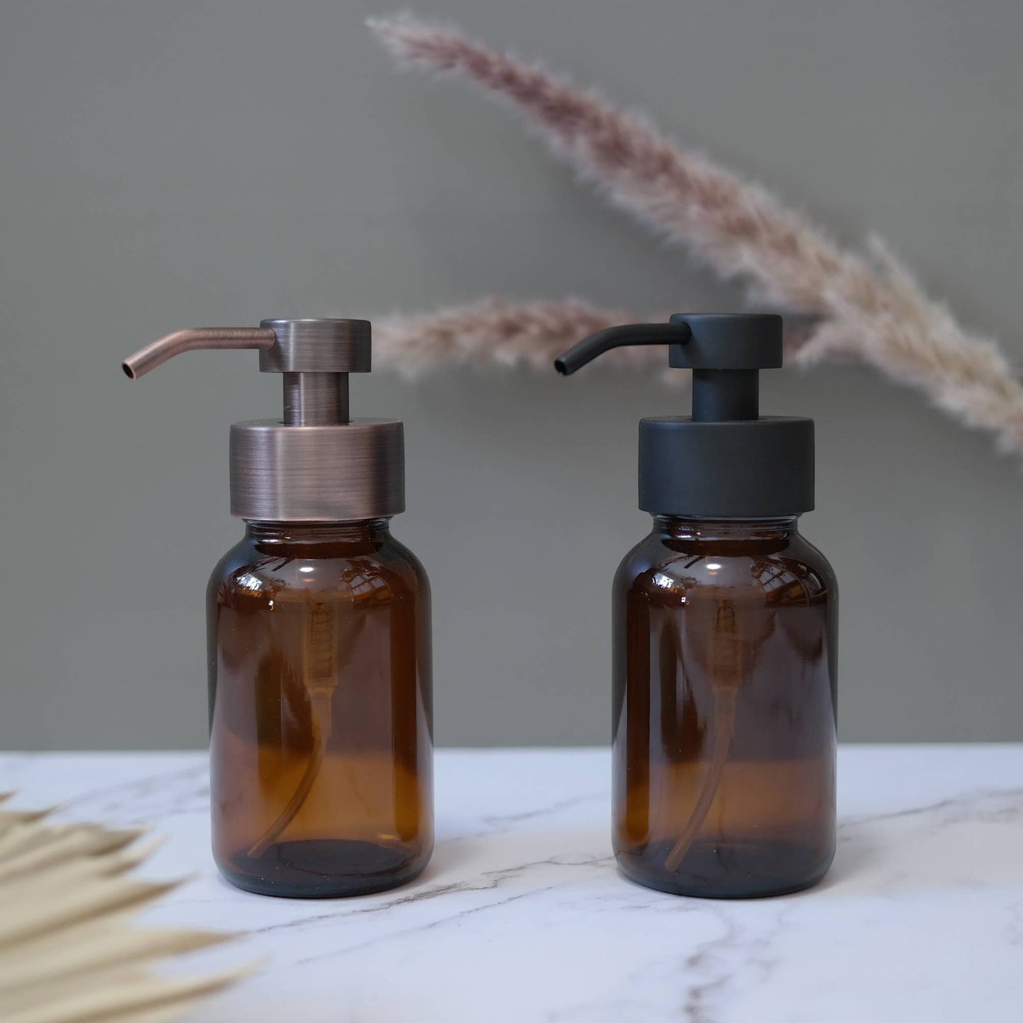 Two brown glass bottles with pump dispensers on a marble surface with dried plants in the background.