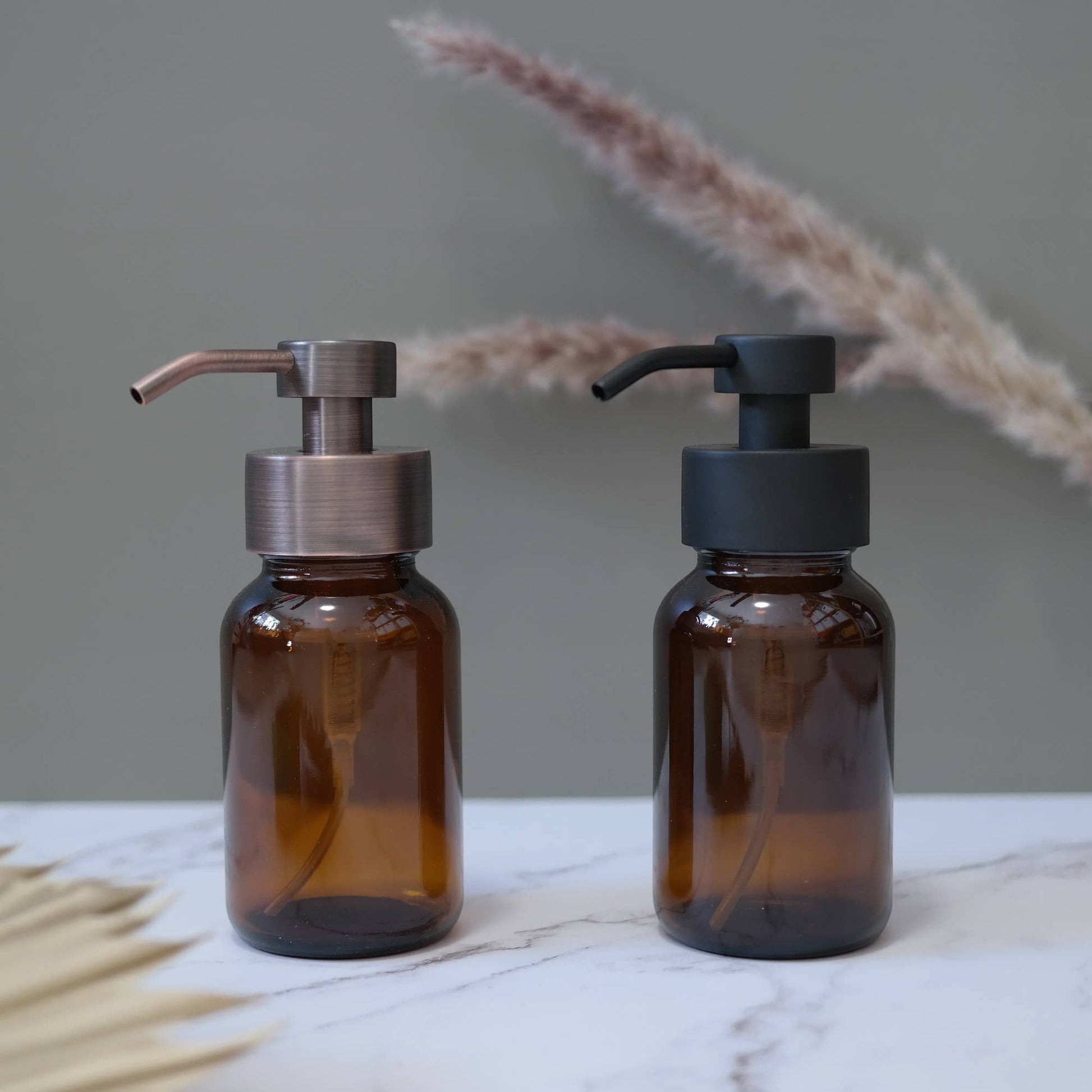 Two brown glass bottles with pump dispensers on a marble surface with dried plants in the background.