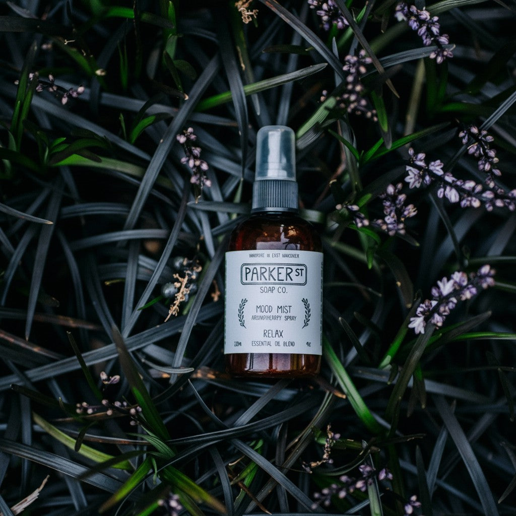 Bubble Gallery RELAX Mood Mist Aromatherapy Spray in amber glass bottle with mister, surrounded by dark foliage and purple flowers.