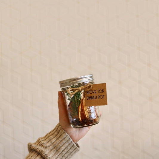 Bubble Gallery spiced simmer pot in a glass jar with dried orange, cinnamon, and evergreen, held by a hand.